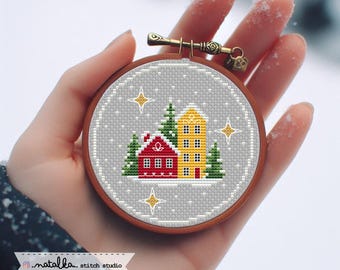 May include: A cross-stitch embroidery hoop featuring a winter scene with a red and yellow house, green trees, and gold stars on a gray background. The wooden hoop is held in a hand. The text "natalka... stitch studio" is at the bottom.