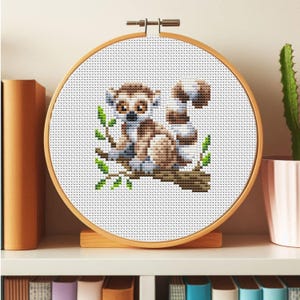 May include: Cross-stitch embroidery hoop with a white background and a colorful image of a lemur sitting on a branch with green leaves.