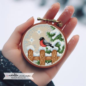 Könnte beinhalten: Ein Kreuzstich-Stickrahmen mit einer Winterszene, die einen rot-schwarzen Vogel auf einem schneebedeckten Zaun zeigt. Das Design umfasst grüne Kiefern und einen hellbeigen Hintergrund mit Sternakzenten. Der Rahmen hat einen Durchmesser von etwa 10 cm.