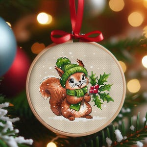 Op de afbeelding: Een kruissteek kerstversiering met een bruin eekhoorntje met een groene muts en sjaal, met hulst met rode bessen. De versiering zit in een houten hoepel, met een rood lint om op te hangen. Sneeuwvlokken zijn verspreid.