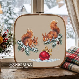 May include: A square wooden embroidery hoop containing a cross-stitch pattern on beige fabric. The design features two brown squirrels perched on snow-covered pine branches with pinecones, and a red Christmas ornament with a gold ribbon below.