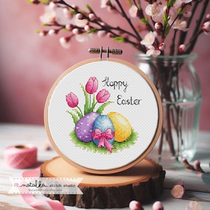 May include: A cross stitch pattern featuring three colorful Easter eggs with a pink bow and three pink tulips. The pattern is framed in a wooden hoop and the text "Happy Easter" is stitched above the eggs.