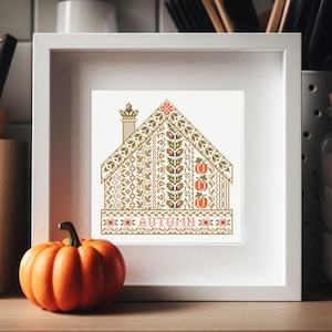 May include: A white frame with a cross-stitch design of a house with a chimney and the word "AUTUMN" underneath. The house is decorated with fall-themed motifs like leaves, pumpkins, and acorns. A small orange pumpkin sits in front of the frame.