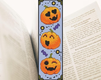 May include: A handmade bookmark featuring three smiling orange pumpkins with black faces and green stems, separated by purple candies, on a light blue background. The bookmark is made with cross-stitch.