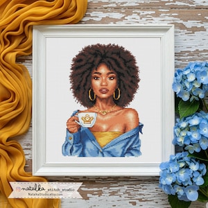 May include: Cross-stitch art of a woman with brown curly hair, gold hoop earrings, and a gold necklace, holding a teacup. She wears a yellow top and a blue denim shirt. The artwork is framed in a white square frame. The background includes yellow fabric and blue flowers.