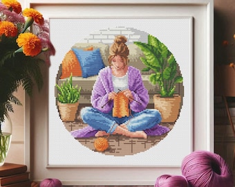 Può includere: Una donna seduta sul pavimento con un maglione viola e jeans blu, a maglia una sciarpa arancione. È circondata da piante e un divano con un cuscino blu e bianco.