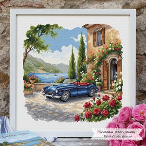 Puede incluir: Obra de punto de cruz enmarcada en blanco. La escena representa un coche descapotable azul aparcado frente a una villa con rosas rojas. El fondo muestra un lago, montañas y árboles. El texto "Dolce Vita" es visible.