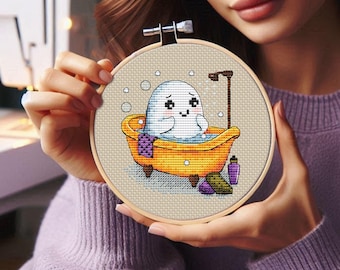 May include: A cross-stitch embroidery hoop with a white ghost taking a bath in a yellow bathtub. The ghost has a happy expression and is surrounded by bubbles. The bathtub has a purple towel and a bottle of soap.
