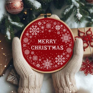 Può includere: Un telaio da ricamo rosso con le parole "MERRY CHRISTMAS" in bianco. Il design presenta fiocchi di neve bianchi attorno al testo. Il telaio è tenuto da una persona che indossa guanti di maglia beige, con un albero di Natale e decorazioni sullo sfondo.