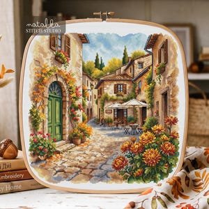 Op de afbeelding: Een kruissteek borduurproject in een houten borduurring met een Europese straatscène met stenen gebouwen, een groene deur, oranje bloemen en cafétafels. De tekst "natalka STITCH STUDIO" staat in de linkerbovenhoek.