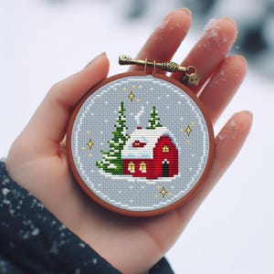 Könnte beinhalten: Ein handgestickter Kreuzstich-Stickrahmen mit einem roten Haus mit weißen Verzierungen und einem schneebedeckten Dach, umgeben von zwei grünen Bäumen und goldenen Sternen auf hellblauem Hintergrund.