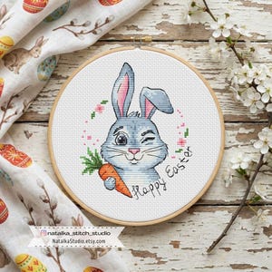 Puede incluir: Un bastidor de bordado de punto de cruz con un conejito gris guiñando un ojo y sosteniendo una zanahoria. El diseño incluye el texto "Happy Easter" y una corona floral. El fondo es blanco y el bastidor es de madera.