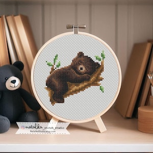 May include: A cross-stitch embroidery of a brown bear resting on a branch, framed in a wooden hoop. The bear is detailed with various shades of brown, and the branch has green leaves. The embroidery is displayed on a wooden stand.