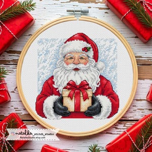 Può includere: Un punto croce di Babbo Natale che tiene un regalo con un fiocco rosso. Babbo Natale indossa un abito rosso con finiture bianche e un cappello abbinato. Lo sfondo è azzurro con un motivo a fiocchi di neve, in un cerchio di legno.