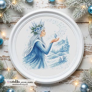 May include: A cross-stitch artwork in a white frame depicts a woman in a blue dress with white hair, holding a crystal. The scene includes a snowy mountain landscape and falling snowflakes. The artwork is framed in a white, round frame.