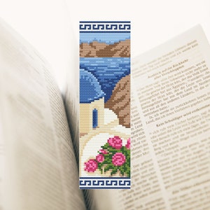 Op de afbeelding: Een kruissteek-bladwijzer met een wit gebouw met een blauwe koepel, omringd door roze bloemen. Het gebouw staat tegen een achtergrond van een blauwe zee en bruine heuvels. De bladwijzer heeft een blauwe en witte Griekse sleutelrand.