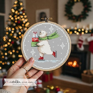 May include: A cross-stitch embroidery hoop featuring a polar bear ice skating and carrying wrapped gifts. The bear wears a green scarf and red ice skates. The background is gray with white snowflake accents. The wooden hoop is held in a hand.