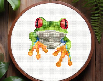 Könnte beinhalten: Kreuzstich-Stickerei eines grünen Laubfrosches mit roten Augen und orangefarbenen Füßen. Der Frosch sitzt auf einem weißen Hintergrund.