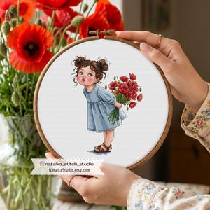 Könnte beinhalten: Ein Kreuzstich-Stickrahmen mit einem jungen Mädchen in einem blauen Kleid, das einen Strauß roter Mohnblumen hält. Die Stickerei ist vor einem weißen Hintergrund. Der Rahmen wird von Händen gehalten. Der Text "natalka_stitch_studio NatalkaStudio.etsy.com" ist sichtbar.