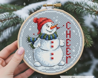 May include: Cross-stitch embroidery of a snowman wearing a red hat and scarf, with the word "CHEER" in red. The snowman has an orange carrot nose and is set in a wooden hoop. The background is a snowy, winter scene.