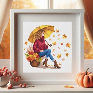 Könnte beinhalten: Eine Frau sitzt unter einem gelben Regenschirm mit einem Korb voller Herbstblätter. Sie trägt einen roten Pullover und eine blaue Jeans. Die Blätter fallen um sie herum.
