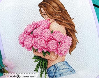 May include: A cross stitch pattern depicting a woman with long brown hair holding a bouquet of pink peonies. The woman is wearing blue jeans and is looking down at the flowers. The pattern is on white Aida cloth.