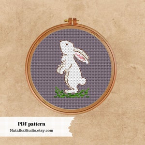 May include: A white rabbit cross stitch pattern in a wooden embroidery hoop. The rabbit is standing on a purple background with green grass.