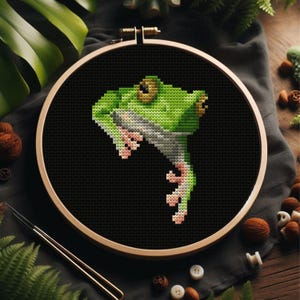 Könnte beinhalten: Kreuzstich-Stickerei einer grünen Frosch auf schwarzem Stoff, in einem Holzreifen. Der Frosch hat detaillierte Merkmale, einschließlich hellgrüner und weißer Schattierungen und rosa Füße. Der Hintergrund ist eine dunkle, einfarbige Farbe.