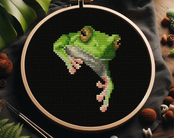 Könnte beinhalten: Kreuzstich-Stickerei einer grünen Frosch auf schwarzem Stoff, in einem Holzreifen. Der Frosch hat detaillierte Merkmale, einschließlich hellgrüner und weißer Schattierungen und rosa Füße. Der Hintergrund ist eine dunkle, einfarbige Farbe.