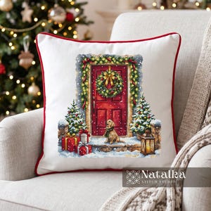 Op de afbeelding: Een wit vierkant kussen met rode bies voorzien van een kruissteekontwerp van een rode voordeur met krans, een golden retriever puppy, twee kleine kerstbomen, ingepakte cadeaus en een lantaarn. De tekst Natalka Stitch Studio is zichtbaar.
