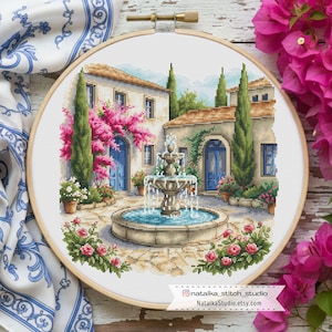 May include: A cross-stitch embroidery of a courtyard scene with a fountain, buildings, and pink flowers. The scene is framed in a wooden embroidery hoop. The buildings have blue doors and windows. The text "natalia_stitch_studio" and "NatalkaStudio.etsy.com" is visible.