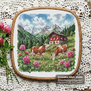 May include: A cross-stitch embroidery in a wooden hoop depicting a mountain landscape with two brown and white cows grazing in a meadow of pink clover flowers, a red house, green pine trees, and snow-capped mountains under a blue sky.