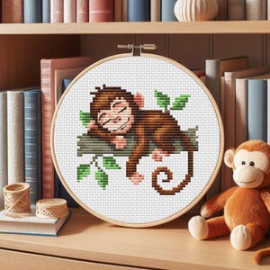 May include: A round embroidery hoop with a white background and a brown monkey sleeping on a branch with green leaves. The monkey has a brown tail and is curled up on the branch.