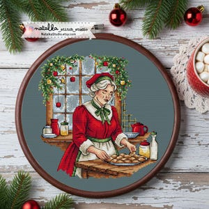 Op de afbeelding: Kruissteekborduurwerk in een houten hoepel toont een vrouw in een rode jurk en groene schort, die koekjes bakt in een kerstversierde keuken. De scène omvat een raam met feestelijke decoraties en de tekst "NatalkaStudio.etsy.com".