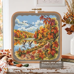 May include: A square wooden embroidery hoop containing a cross-stitch pattern of an autumn landscape with a lake, flying geese, and vibrant orange and red maple leaves. The text natalka_stitch_studio and natalkaStudio.etsy.com is visible.