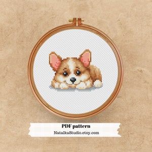 Puede incluir: Patrón de punto de cruz de un perro corgi marrón y blanco con orejas rosas, tumbado y mirando al espectador. El patrón está en un aro de bordado de madera sobre un fondo marrón.