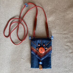Peut inclure: Une petite pochette en tissu bleu avec un motif de perles rouges, orange et brun. La pochette est munie d'un cordon rouge et est décorée d'un gros bouton orange.