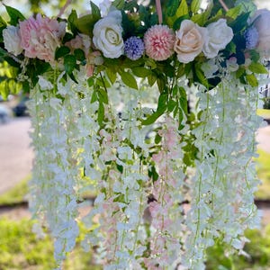 May include: A floral chandelier featuring a ring of white, pink, and purple flowers and greenery. White and pink wisteria-like flowers cascade downwards, suspended by a pink rope. The arrangement is set against a blurred outdoor background.