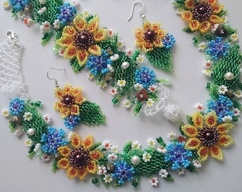 May include: A beaded jewelry set featuring a necklace and matching earrings crafted with intricate glass beads. The design showcases yellow sunflowers with brown centers, blue cornflowers, white daisies, and green leaves, all connected by clear beads.