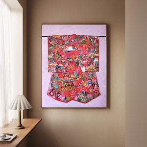 Puede incluir: Una obra de arte enmarcada que presenta un kimono rojo vibrante con un patrón colorido y detallado de pájaros, flores y otros elementos naturales. La obra de arte se exhibe sobre un fondo rosa claro.