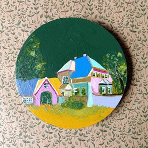 Könnte beinhalten: Ein farbenfrohes Acrylgemälde einer malerischen Dorflandschaft auf einer runden Leinwand. Das Kunstwerk zeigt Häuser mit rosa, blauen und gelben Dächern vor einem dunkelgrünen Hintergrund. Das Gemälde befindet sich auf einem beigefarbenen Hintergrund mit Blättermuster.