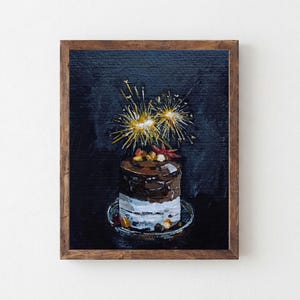 Könnte beinhalten: Ein Ölgemälde in einem braunen Holzrahmen, das einen kleinen Schichtkuchen auf einem Teller vor dunklem Hintergrund zeigt. Der Kuchen ist mit zwei hellgelben Wunderkerzen gekrönt, die Licht ausstrahlen, sowie kleinen roten und braunen Akzenten.