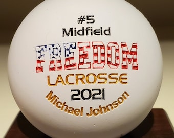 May include: A white lacrosse ball with the words "FREEDOM LACROSSE 2021" in red, white, and blue lettering. The ball also features the name "Michael Johnson" and the number "#5 Midfield".
