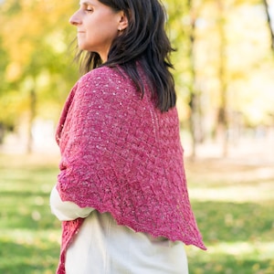 Puede incluir: Una mujer lleva una chal de punto rosa con un patrón de encaje. El chal está drapeado sobre sus hombros y le llega hasta la cintura. Está de pie en un parque con árboles en el fondo.