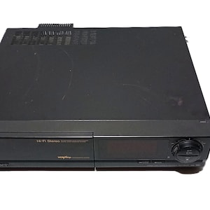 May include: Black Sanyo Hi-Fi Stereo VHS player with a remote control and a Fuji HQ 120 VHS tape. The VCR has a digital display and a control knob. The VHS tape is in its original packaging.