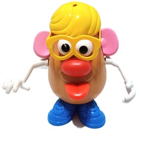 May include: A plastic potato-shaped toy figure with a tan body, yellow hair, and yellow glasses. It features pink ears, a red nose, a red tongue, white arms with hands, and blue shoes. The figure has large white eyes with black pupils.