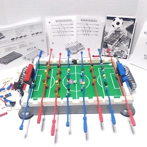 May include: A miniature foosball table with a green playing field, red and blue player rods, and a small soccer ball. The set includes instructions and unassembled parts. The table has a white border and black nets at each end.