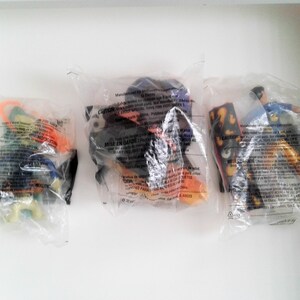 May include: Three plastic bags containing small toys. The toys are in various colours, including orange, yellow, blue, green, and black. The bags have warning labels in English and French.