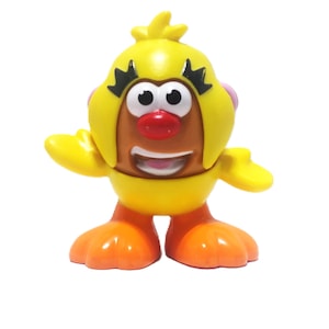 May include: A yellow and orange toy figure with a cartoonish design. The figure has a yellow head with black accents, a red nose, and a smiling mouth. The figure's arms and feet are orange.