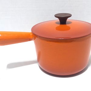 May include: An orange enameled cast iron saucepan with a lid. The saucepan has a single handle and a dark brown knob on the lid. The cookware is a vibrant orange color.
